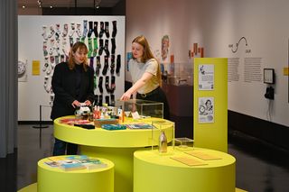 Zwei Besucherinnen stehen an einer Gruppe von gelben Tischen in verschiedener Höhe, auf denen verschiedenste Objekte ausliegen, teilweise unter Glaskästen. Im Hintergrund hängen bunte Socken paarweise an einer weißen Wand.
