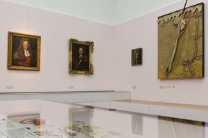 Vpravo na stěně model vlasového kořene a nalevo tři historické portréty osob s parukami.