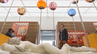 Mladý muž před modelem hermafroditu pozoruje barevné kuličky na tyčkách rozmístěné v sálu. 