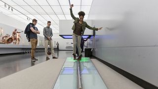 Mladá žena balancuje na kovovém chodníku pro test rovnováhy v bílém sálu Pohyb. Dva mladí muži ji při tom sledují.