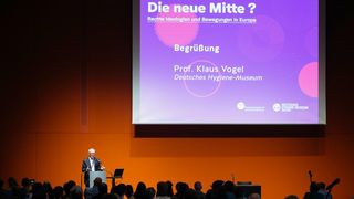 Der Direktor des Museums, Professor Klaus Vogel, am Stehpult vor einer violetten Hintergrundpräsentation.