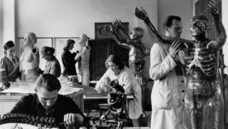 Stará černobílá fotografie zaměstnanců dílen muzea při práci na skleněných figurách. Dva z nich v bílých pláštích.
