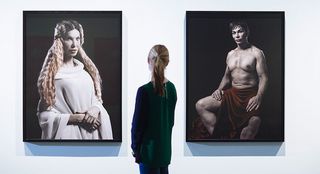 Eine junge Frau vor einer weißen Wand zwischen zwei großformatigen Fotografien. Sie zeigen eine blonde Frau in weißem Kleid und einen braunhaarigen halbnackten Mann, die sich Schönheitsoperationen unterzogen haben.