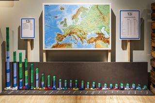 Auf einer hölzernen Versandkiste stehen 25 Spielzeugautos in einer Schlange. Auf den Spielzeugautos sind Bauklötze in unterschiedlicher Höhe befestigt, die für die Herkunftsländern von Ausländern und Deutschen mit Migrationshintergrund in Deutschland stehen. Der größte Stapel hat die Höhe von einer Armlänge, die anderen Stapel werden gestaffelt kleiner. Dahinter eine Landkarte von Europa.