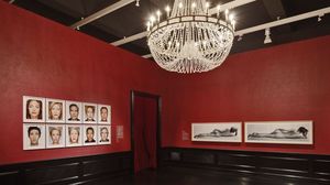 Ein Austellungsraum mit roten Wänden. An der Decke hängt ein pompöser Kronleuchter. An der Wand hängen zehn Portäts prominter Personen.