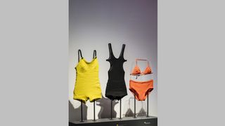 Vor einer weißen Wand steht ein quaderförmiger Sockel. Auf ihn ein gelber Badeanzug, ein schwarzer Badeanzug und ein roter Bikini. Alle drei sind ältere Modelle.