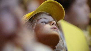 Ein Mädchen aus der Froschperspektive gezeigt, mit gelben Cappy. Sie schaut nach rechts. Die Personen um sie herum sind verschwommen.