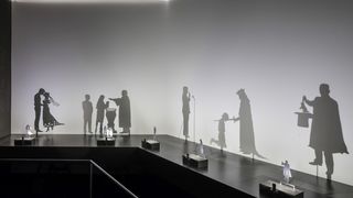 Das Schattentheater vor einer weißen Wand, das mit kleinen animierten Silhouetten auf einem schwarzen Stand Sprachrituale, zum Beispiel bei der Hochzeit oder der Taufe, nachstellt.