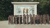 Eine große Gruppe Frauen in militärischer Uniform mit Schirmmützen steht vor einem Wandmosaik. Darauf ist eine Frau mit einem kleinen Jungen auf der Schulter zu sehen sowie ein Mann, der die Hand des Jungen hält. Alle tragen Uniformen. Hinter der Familie ist eine kleine Blockhütte sowie ein schneebedeckter, bewaldeter Berg zu sehen. Hinter dem Mosaik sieht man ebenfalls Wald.