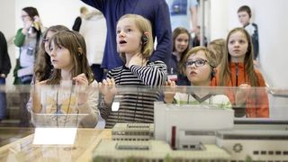 Kinder mit Kopfhörern vor einer Vitrine, von vorn fotografiert, mit Blick nach oben. In der Vitrine ein Modell in Miniatur des Hygiene-Museums.