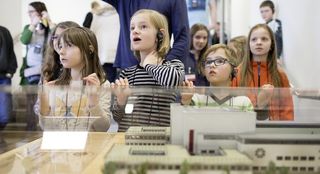 Kinder mit Kopfhörern vor einer Vitrine, von vorn fotografiert, mit Blick nach oben. In der Vitrine ein Modell in Miniatur des Hygiene-Museums.