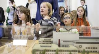 Kinder mit Kopfhörern vor einer Vitrine, von vorn fotografiert, mit Blick nach oben. In der Vitrine ein Modell in Miniatur des Hygiene-Museums.