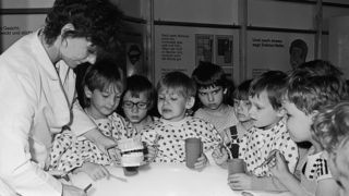 Bild aus der DDR-Zeit: Eine Mitarbeiterin demonstriert einer Gruppe von Kindern mit Zahnmodell, Zahnputzbecher und Zahnbürste, wie man richtig Zähne putzt. 