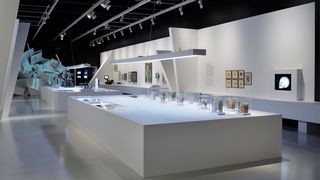 Ein Ausstellungsraum mit weißen Wänden. In der Mitte des Raumes ein breiter und langer weißer Block auf den Vitrinen mit Exponaten stehen.