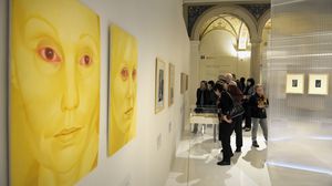 An der Wand eines Ausstellungsraumes hängen zwei Gemälde. Dargestellt sind zwei Frauen, deren Augen rot sind. Der restliche Hintergrund und die Frauen selbst sind gelb.
