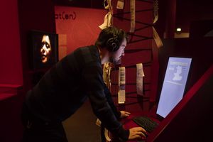 Ein Mann mit Kopfhörern steht an einem Computer in der Ausstellung Sprache. Der Hintergrund ist abgedunkelt und die Wände sind rot.
