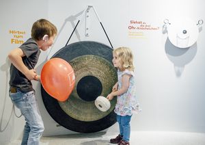 Ein Mädchen steht vor einer Gongschale, die so groß ist wie sie. Links im Bild steht ein Junge mit einem roten Luftballon in der Hand.