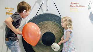 Ein Mädchen steht vor einer Gongschale, die so groß ist wie sie. Links im Bild steht ein Junge mit einem roten Luftballon in der Hand.