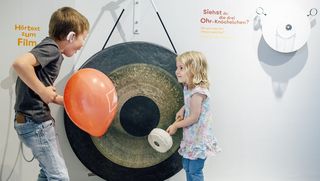 Ein Mädchen steht vor einer Gongschale, die so groß ist wie sie. Links im Bild steht ein Junge mit einem roten Luftballon in der Hand.