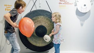 Ein Mädchen steht vor einer Gongschale, die so groß ist wie sie. Links im Bild steht ein Junge mit einem roten Luftballon in der Hand.