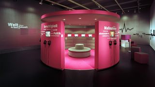 Ein großer pinker zylinderförmiger Pavillion steht in der Raummitte. Im Hintergrund sind schwarze Wände auf denen Grafiken oder Worte stehen. Rechts sind außerdem Medienstationen.