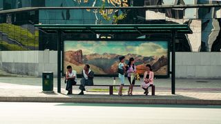 Eine Bushaltestelle in Pjöngjang, Nordkorea. Zwei Männer und drei Frauen warten auf den Bus. Die Männer tragen Anzughosen und weiße Hemden, die Frauen tragen Kostüm in Pastellfarben. An der Rückwand der Bushaltestelle ist ein großes Bergpanorama zu sehen. Im Hintergrund befindet sich ein großes, modernes Gebäude.