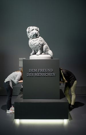 Vor einer dunkelgrauen Wand befindet sich die weiße Skulptur eines langhaarigen Hundes. Diese Skulptur steht auf einem Sockel mit der Aufschrift "Dem Freund Des Menschen".