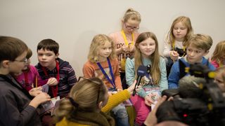 Eine Reporterin hält einem Mädchen ein Mikrophon für ein Interview entgegen. Neben dem Mädchen sitzen noch sieben weitere Kinder.