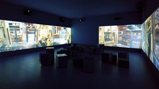 In einem dunklen Ausstellungsraum werden an die linke, rechte und hintere Wand Filmausschnitte projiziert. In den Filmausschnitten unterschiedliche Aufnahmen eines Lagerhalle.