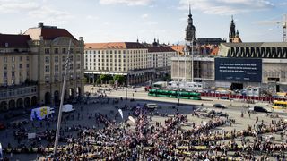 Sehr viele Menschen stehen oder sitzen strahlenförmig an langen gelben Picknicktischen draußen in der Sonne auf dem Altmarkt von Dresden.