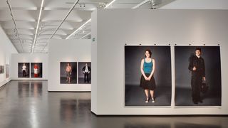 Ein Ausstellungsraum an dessen Wänden drei Bilder hängen, auf denen lebensgroß Menschen abgebildet sind. Auf dem vordergründigsten Bild die zwei Fotos einer Frau. Auf dem einen Foto trägt sie eine Schornsteinfeger-Uniform. Auf dem anderen Bild trägt sie einen schwarzen Rock und ein dunkelgrünes Oberteil.