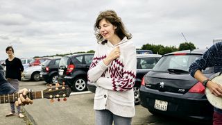 Eine christliche Festivalbesucherin in einer weißgemusterten Strickjacke auf einem Parkplatz. Ihre Augen sind geschlossen, links und rechts von ihr spielen Leute Musik.