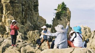 Vier Frauen in Wanderkleidung und Sonnenhüten posieren auf Felsen für ein Foto. Eine fünfte steht ein Stück abseits mit einem Smartphone und fotografiert sie. Im Hinergrund sind steile Felsformationen mit einzelnen Nadelbäumen sowie ein weiterer Wanderer.