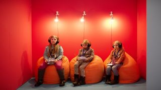 Drei Mädchen sitzen vor einer roten Wand auf orangen Sitzsäcken und haben Kopfhörer auf.