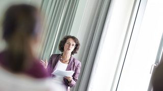 Eine braunhaarige Frau mit Brille und Papieren in der Hand spricht vor einer Gruppe.