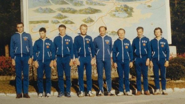 Acht Europäer stehen nebeneinander vor einer großen Wanderkarte mit koreanischer Beschriftung. Die Männer tragen identische blaue Sportanzüge, die über der linken Brust den Schriftug "DDR" und das Staatswappen, Hammer und Zirkel im Ährenkranz, zeigen.