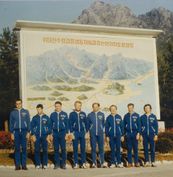 Acht Europäer stehen nebeneinander vor einer großen Wanderkarte mit koreanischer Beschriftung. Die Männer tragen identische blaue Sportanzüge, die über der linken Brust den Schriftug &quot;DDR&quot; und das Staatswappen, Hammer und Zirkel im Ährenkranz, zeigen.