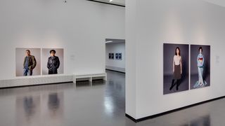 Ein Ausstellungraum an dessen Wänden zwei Bilder hängen, auf denen lebensgroß Menschen abgebildet sind. Auf dem einen Bild eine Frau, die ein Gesha-Kostüm trägt und daneben einen Rock mit T-Shrit. Auf dem anderen Bild ein Mann, der eine Polizei-Uniform trägt und daneben eine Jeans, ein gestreiftes Oberteil und eine kakifarbene Jacke.