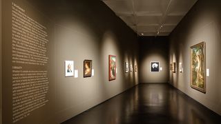 Ausstellungsraum mit Gemälden und Fotografien an dunklen Wänden. Links im Vordergrund steht ein Ausstellungstext an der Wand.