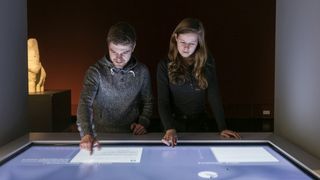 Eine junge Frau und ein junger Mann an einem Touchscreen-Tisch in einem dunklen Raum.