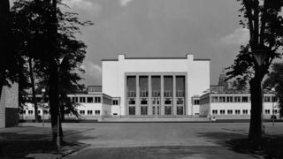 Schwarzweißes Foto im Hintergrund mit dem Museum von vorn. Im Vordergrund stehen links und rechts Bäume.