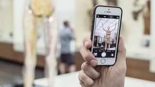 Im Vordergrund eine Hand mit einem Smart-Phone. Das Display zeigt die die Fotofunktion. Die Gläserne Frau wird gerade fotografiert.