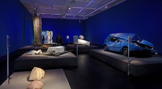 Ein dunkelblauer Ausstellungsraum. Im Vordergrund zwei Felsstücke, rechts ein verbeultes, blaues Auto. Im Hintergrund eine zerstörtes Straßenschild und ein Baum mit Blitzschlag.