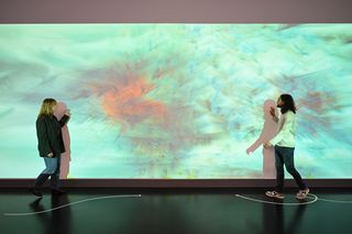 Eine junge Frau und ein junger Mann gehen vor der Projektion im Foyer aufeinander zu und streichen mit den Händen über die Projektionsfläche. Die Projektion zeigt ein abstraktes Muster mit bunten Farben vor einem bläulichen Hintergrund.