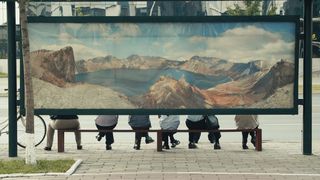 An der Rückwand einer Bushaltestelle in Nordkorea ist ein großes Panoramabild angebracht, dass einen tiefblauen Bergsee umgeben von Berggipfeln zeigt. Unterhalb des Bildes sind von der Hüfte abwärts Personen zu sehen, die auf einer Bank sitzen und auf den Bus warten.