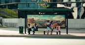 Eine Bushaltestelle in Pjöngjang, Nordkorea. Zwei Männer und drei Frauen warten auf den Bus. Die Männer tragen Anzughosen und weiße Hemden, die Frauen tragen Kostüm in Pastellfarben. An der Rückwand der Bushaltestelle ist ein großes Bergpanorama zu sehen. Im Hintergrund befindet sich ein großes, modernes Gebäude.