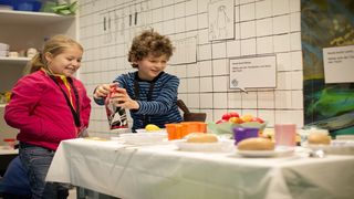 Ein Junge und ein Mädchen stehen an einem Tisch mit Plastik-Lebensmitteln und betrachten diese.