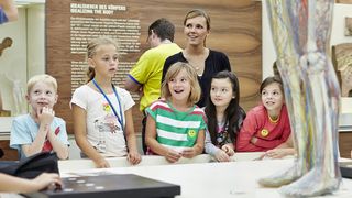 Eine Gruppe sommerlich-bunt gekleideter Kinder und ihre Betreuerin versammeln sich um die interaktive Gläsernen Frau.