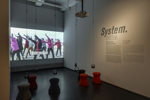 Ein Raum an dessen hinterer Wand eine Leinwand hängt, auf die ein Film projiziert wird. Im Film tanzen 10 Menschen syncron. Davor fünf Hocker und Kopfhörer, die von der Decke hängen.