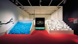 Auf der rechten Seite eines Ausstellungsraumes eine weiße Treppe, auf deren Stufen Sportschuhe in einer Reihe stehen. Auf der linken Seite ein Stapel mit hellblauen Plastikteilen.